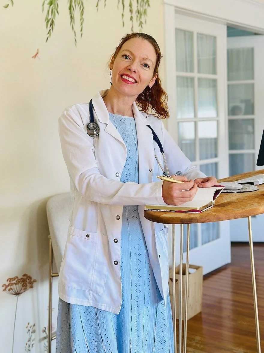 Dr. Abigail Stephens smiling in a white coat with a stethoscope, writing in a journal at a standing desk in a sunny room with a willow mural at Wildflower Integrative Medicine.