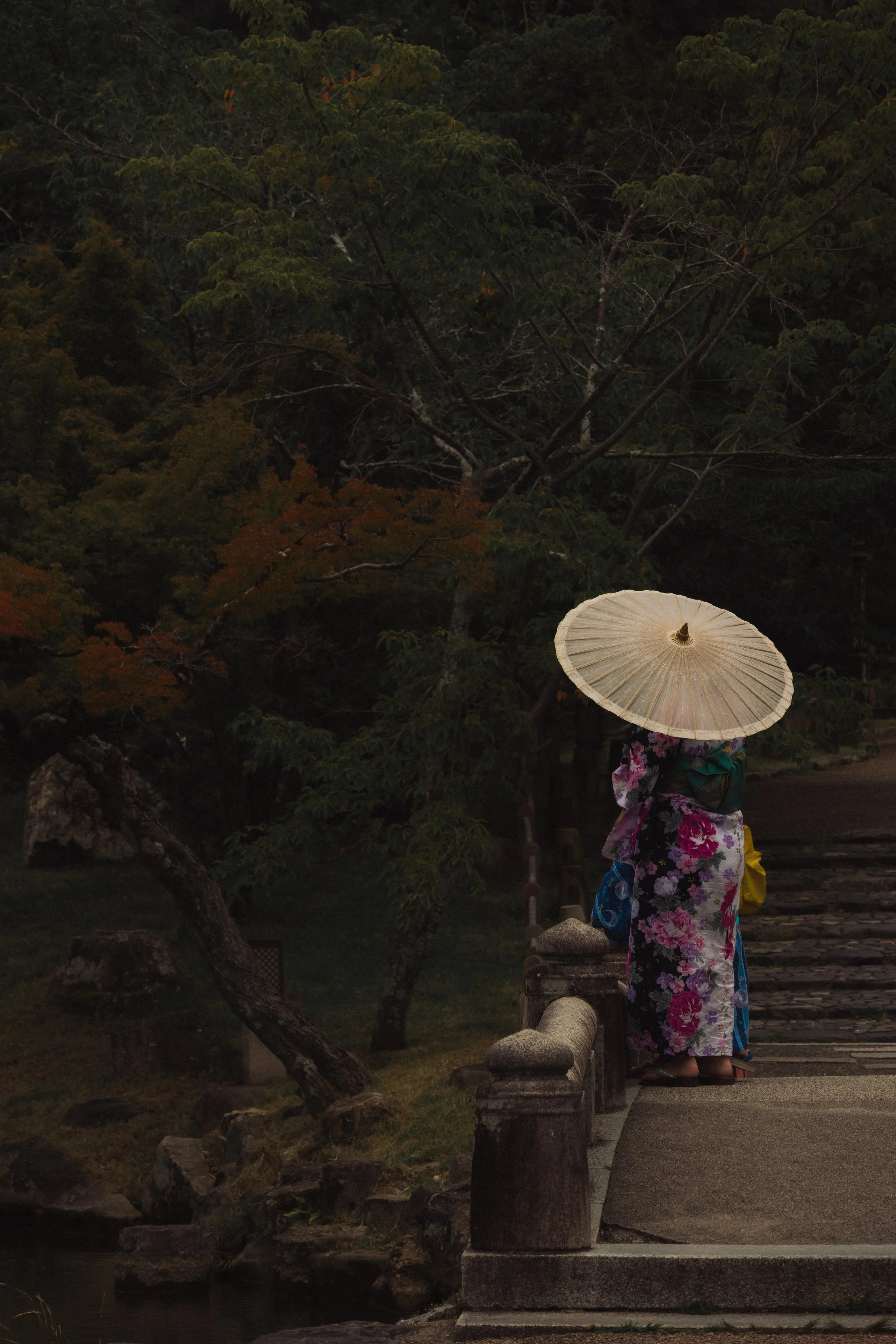 A figure in yukata with a paper parasol on a stone bridge in a Japanese garden