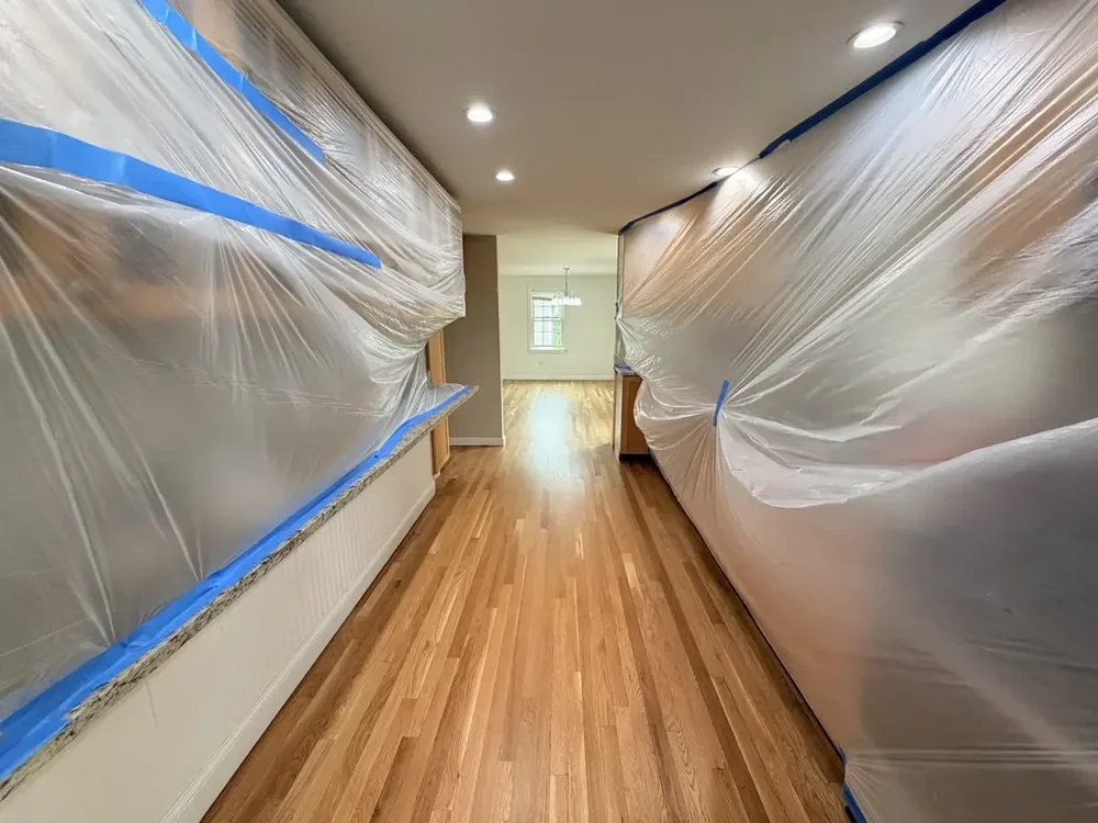 Cabinetry on both sides of a room covered with plastic sheeting secured with blue tape.