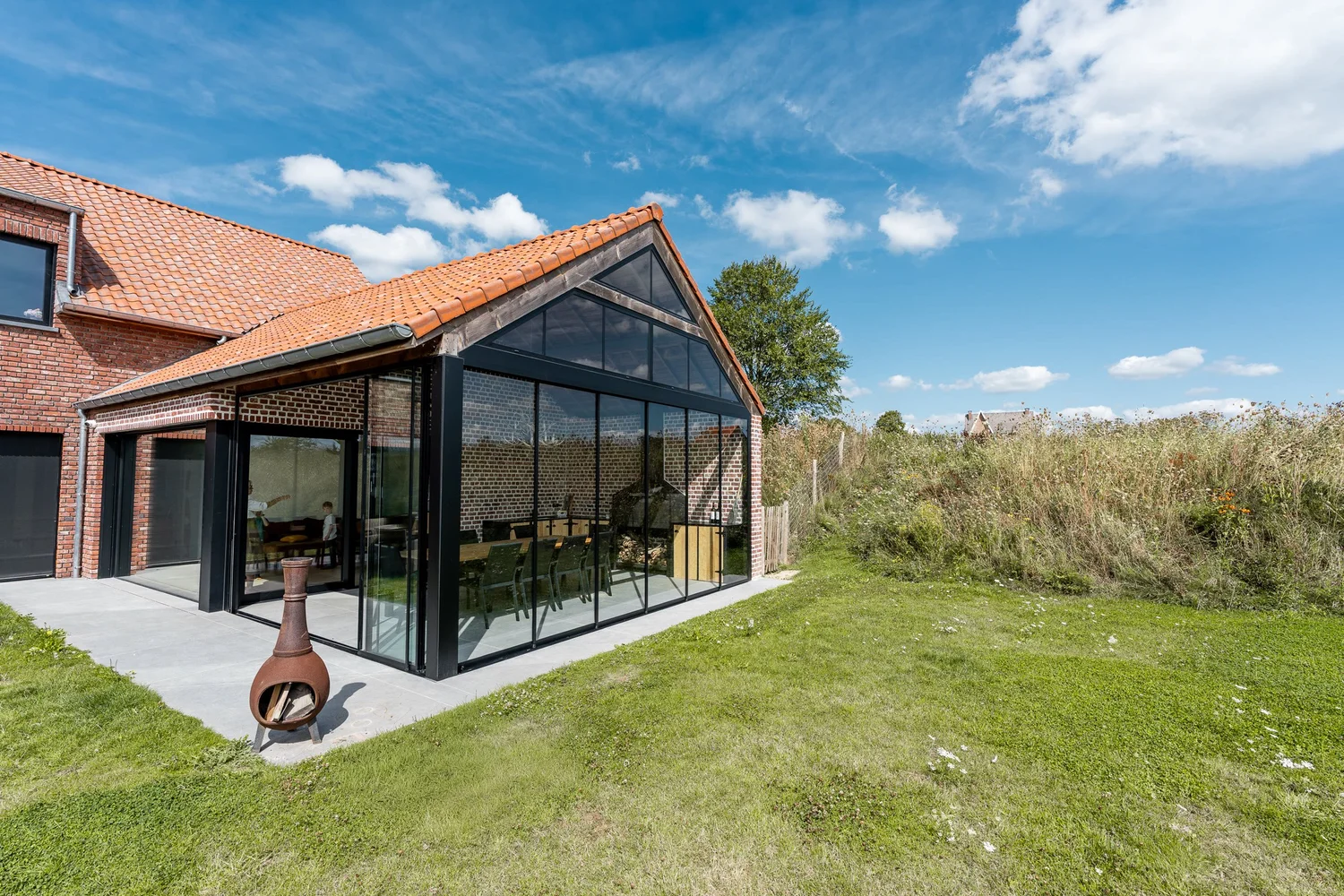 Glazen buitenruimte met glazen geveltop tegen een bakstenen woning in Lennik, afgesloten met omkaderde glazen schuifwanden