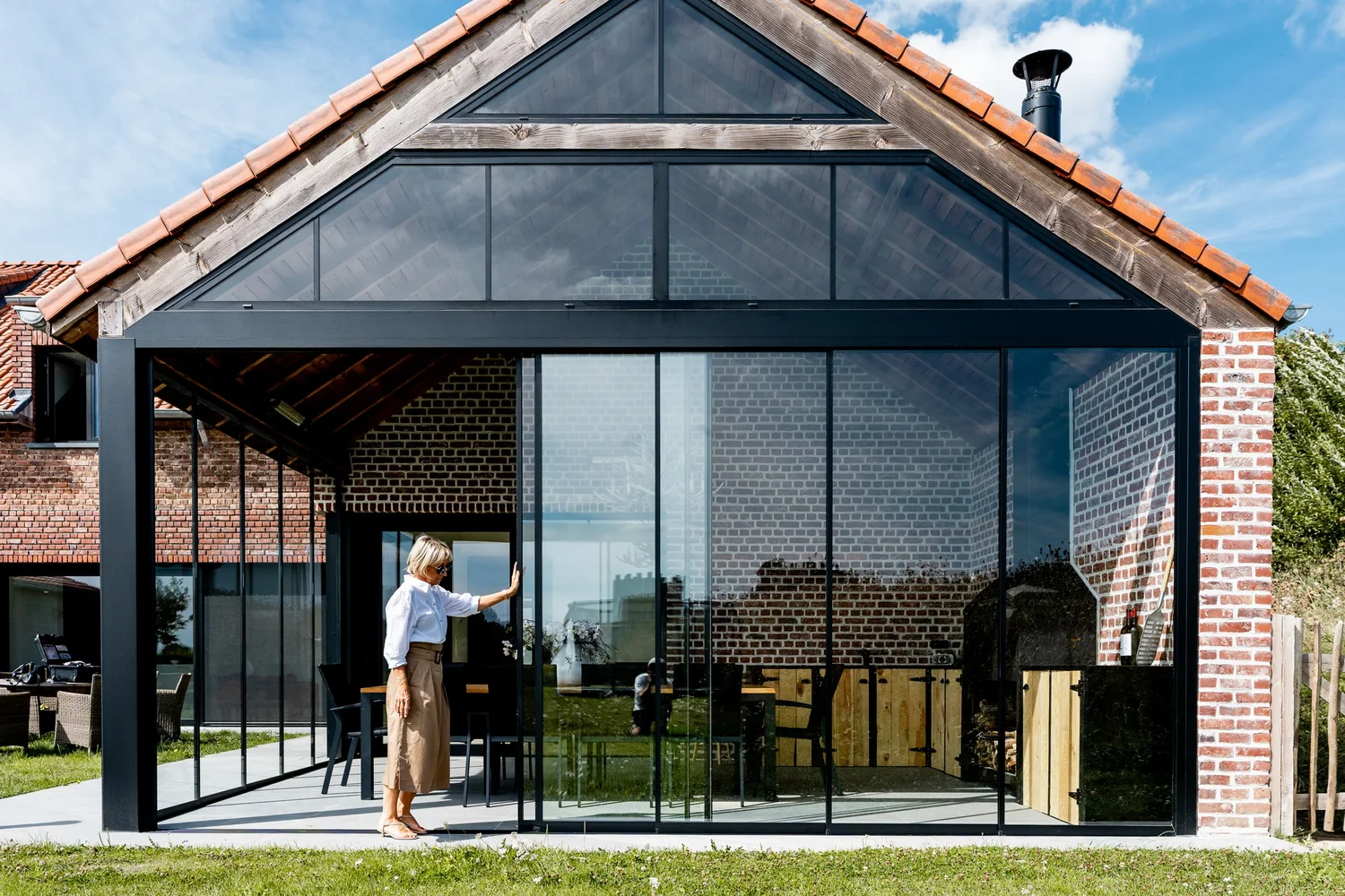 Glazen geveltop en omkaderde schuifwanden aan een bakstenen woning in Lennik, een vrouw schuift de wand open
