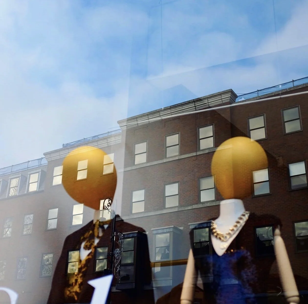 Yellow-headed mannequins behind glass overlaid by reflections of buildings and blue sky