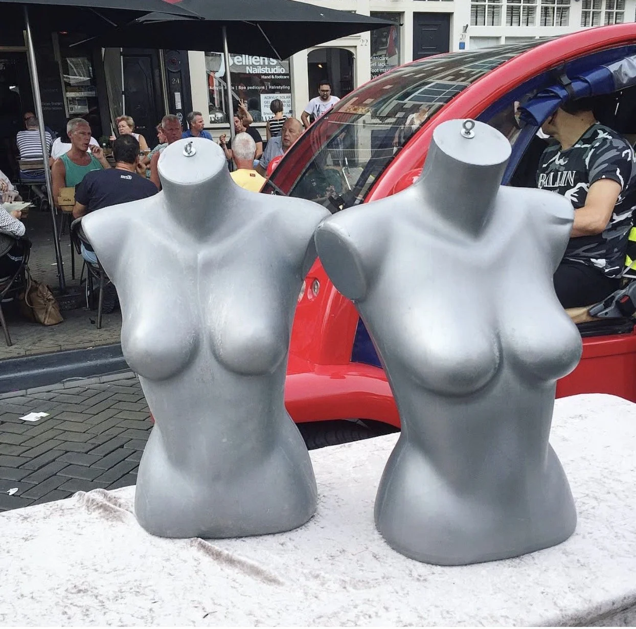 Two silver mannequin torsos beside a red vehicle and people sitting outside