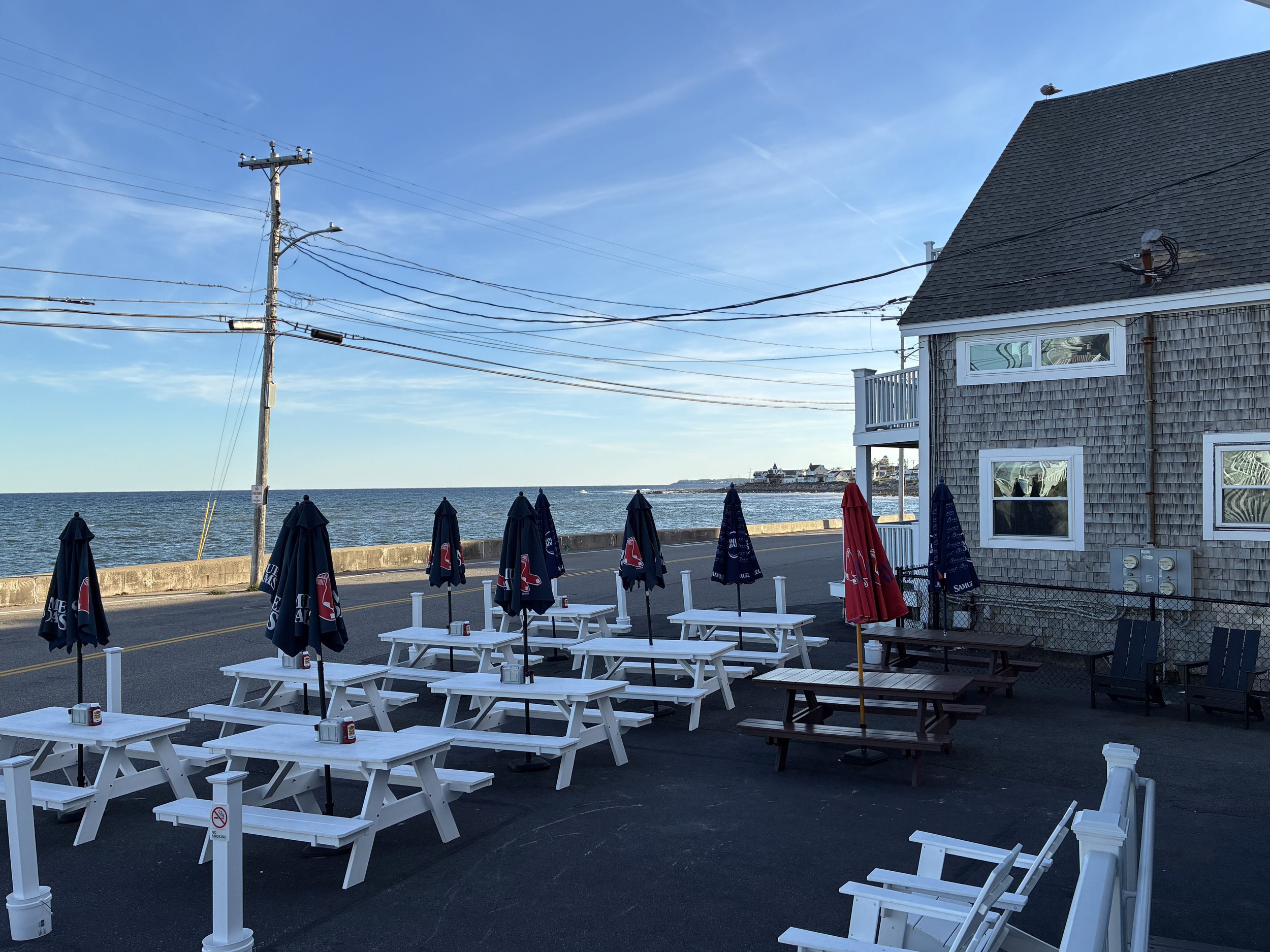 An oceanfront restaurant patio with picnic tables beside a shingled building.