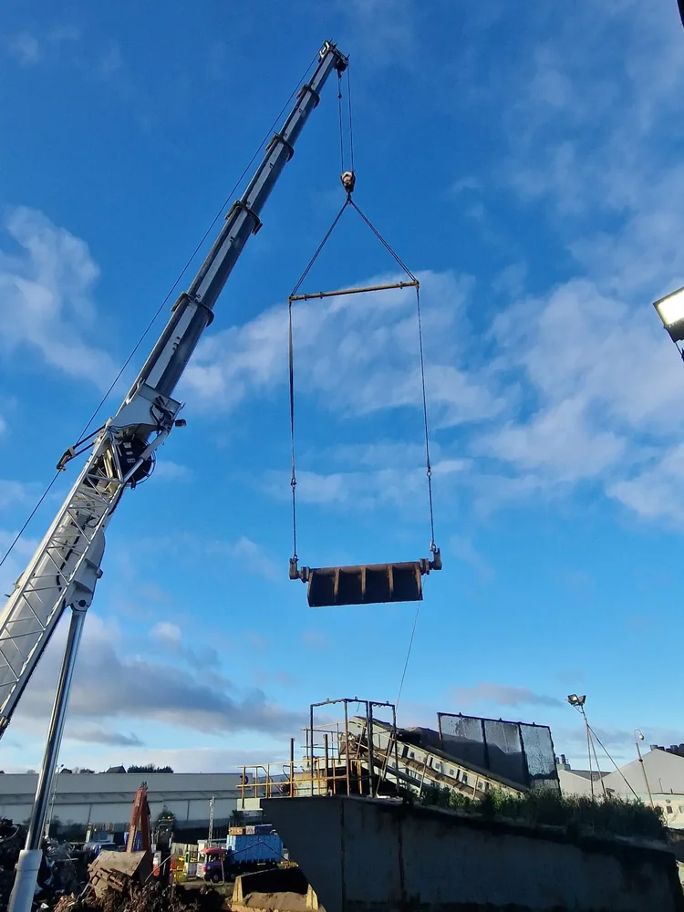 A crane lifting a large machine component using a spreader beam above a processing site