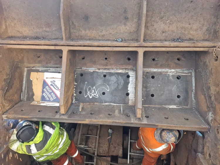 Two workers carrying out repair work beneath a steel machine housing with welded replacement plates