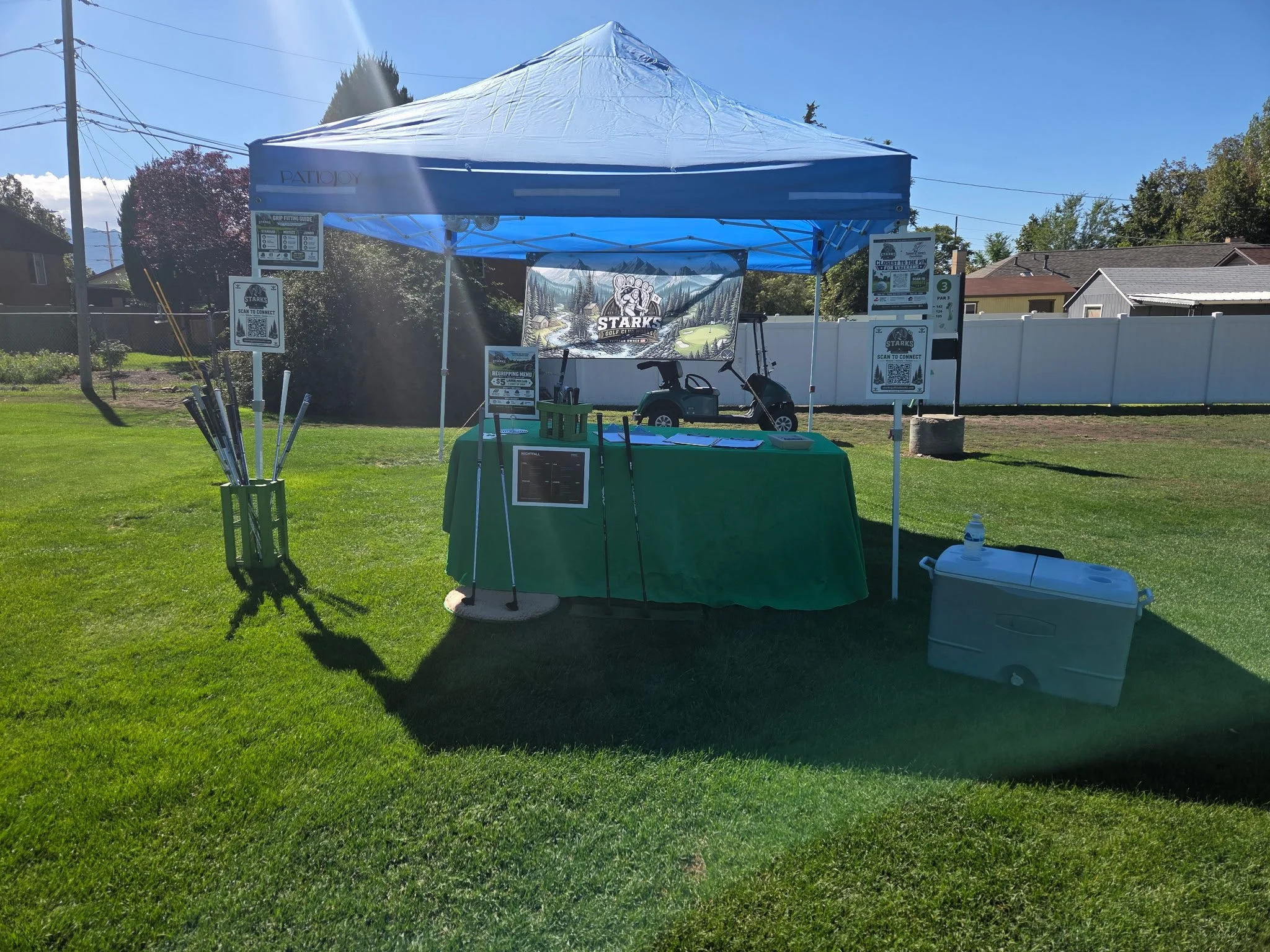 Wide view of the Starks Golf Club Works booth with a blue canopy, green table, grip displays and fundraiser signage at Forest Dale Hole #3.