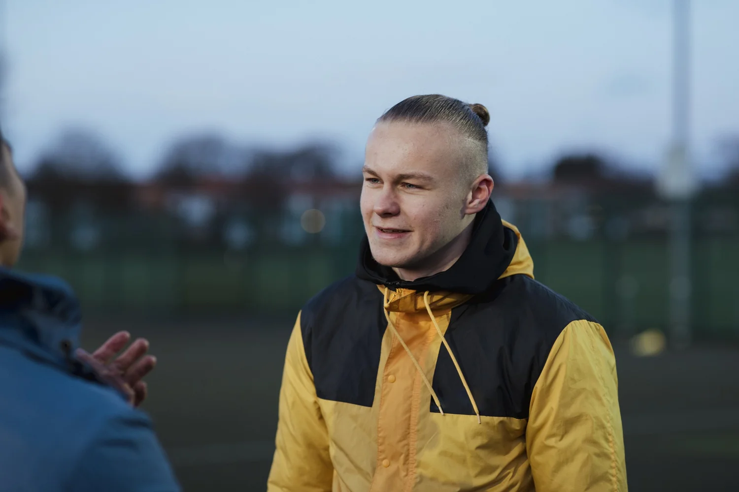 A teenage footballer wearing a yellow and black jacket on a sports field