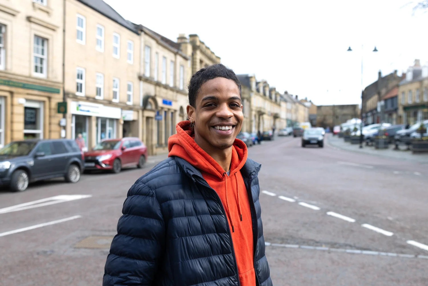 A smiling young man wearing a dark jacket over an orange hoodie outdoors