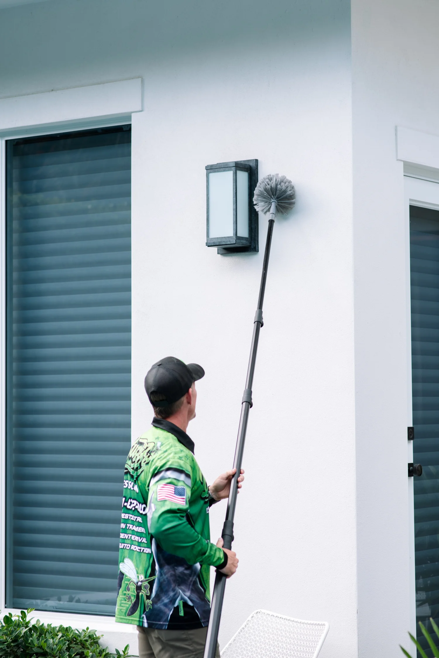 Buggify technician reaching a pole duster up to a wall light beside shutters