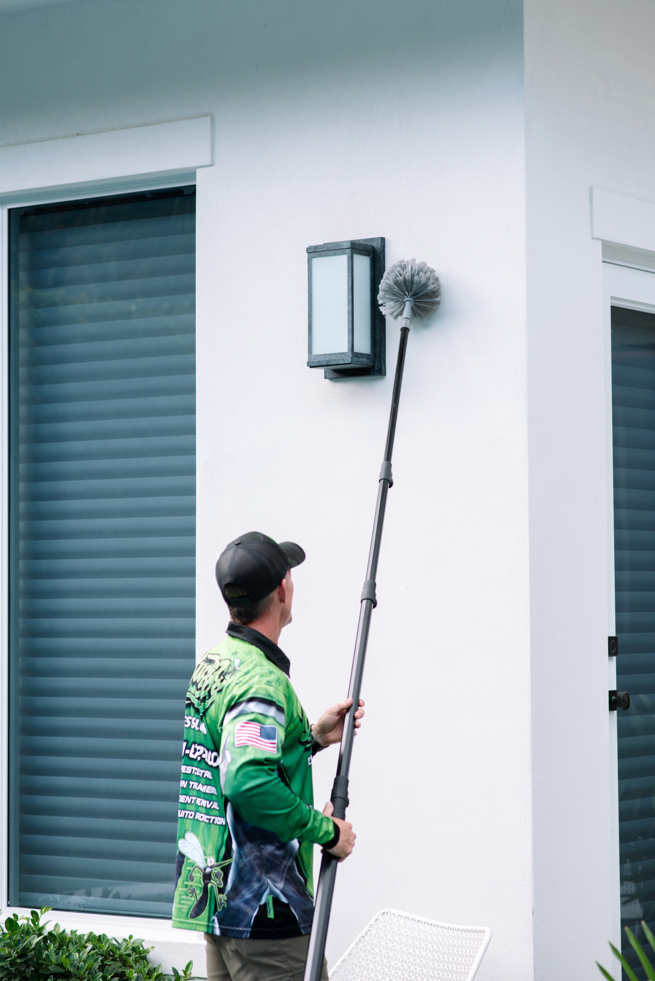 Buggify technician reaching a pole duster up to a wall light beside shutters