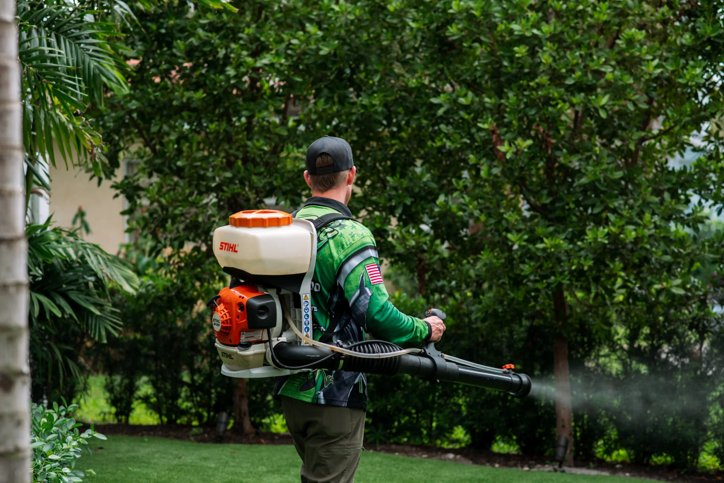 Buggify technician with a mist-blower spraying along a hedge line, mist visible, shown faintly behind the FAQ