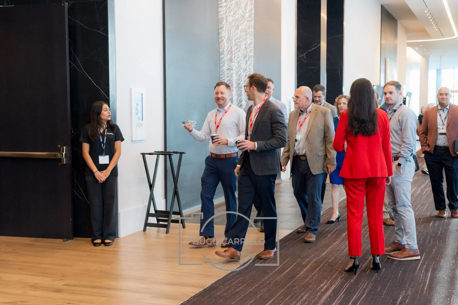 A group of business professionals walking in a conference or event hallway, some holding drinks, with a woman in red pants and blazer and a man in a grey blazer and glasses at the front.