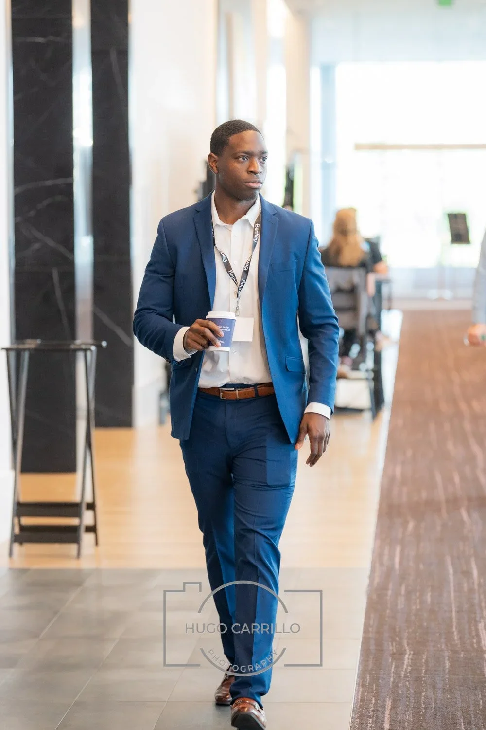 A man in a blue suit and white shirt walking through a conference or office lobby, holding a coffee cup.