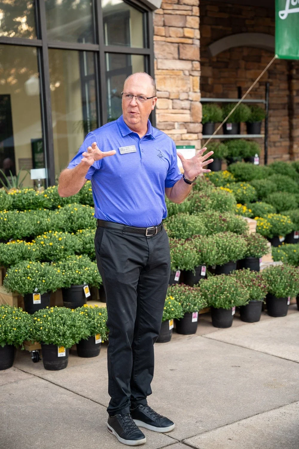 A man wearing glasses, a blue polo shirt, and black pants is standing outside a garden center with potted plants behind him, speaking or giving a presentation.