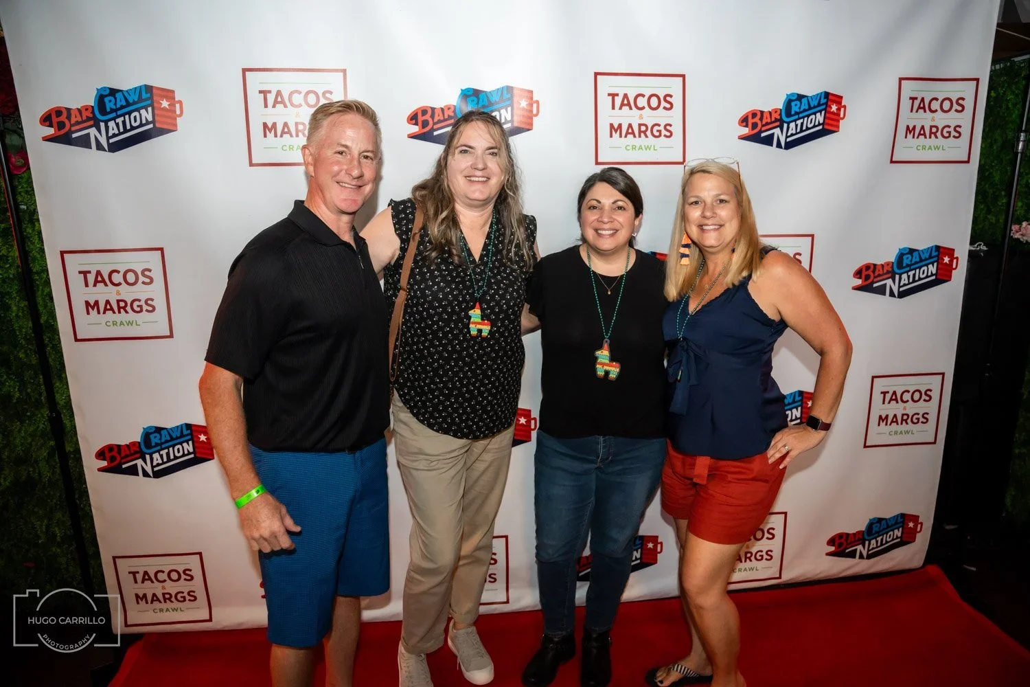 Four people standing in front of a step-and-repeat banner that features logos for 'Bar Rawl Nation' and 'Tacos & Margs Crawl.' They are smiling and wearing colorful necklaces and casual summer clothing.
