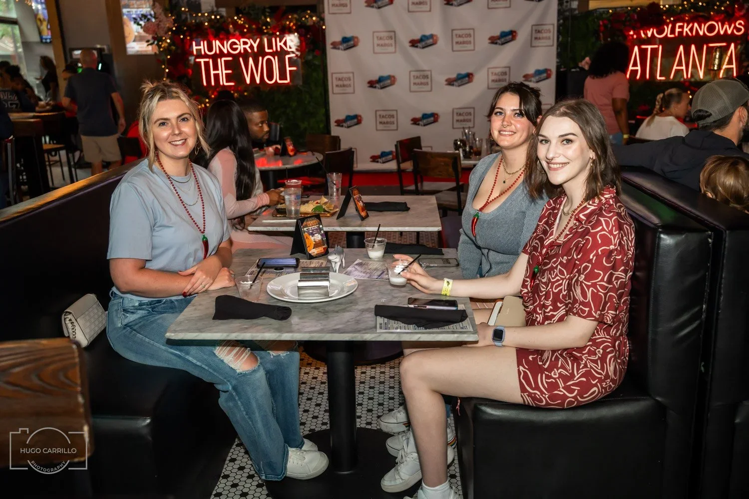 Three young women sitting at a restaurant table smiling at the camera, with neon signs in the background that say 'Hungry Like the Wolf' and 'Wolfknows Atlanta'.