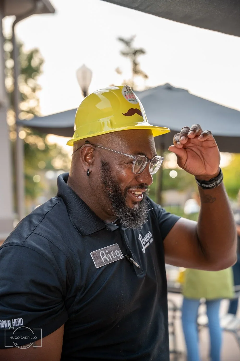 A smiling man wearing glasses, a black shirt with a name tag that says 'Rico,' and a yellow hard hat with a mustache and sticker, outdoors at a restaurant or cafe with umbrellas in the background.