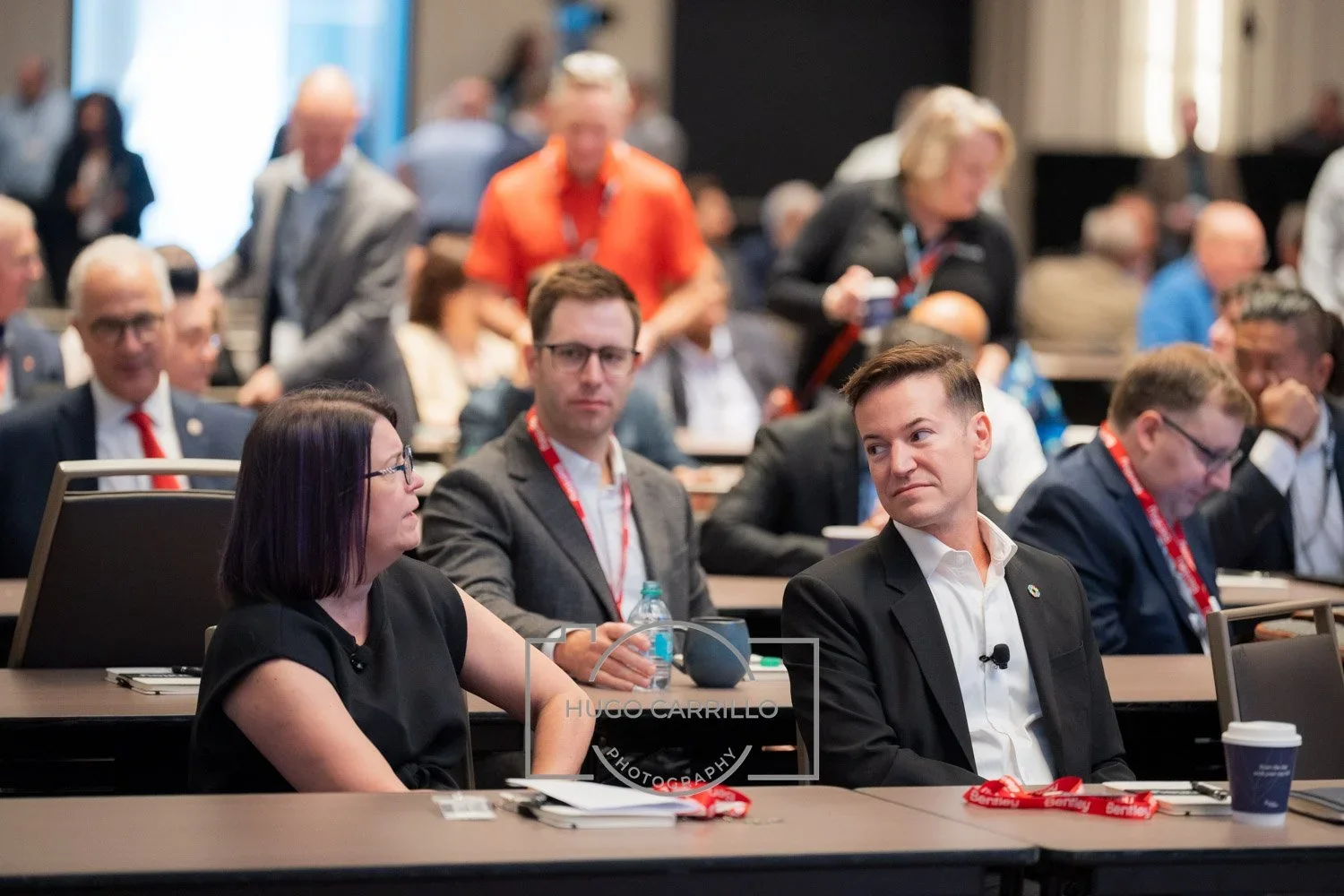 Attendees participate in a conference or seminar, sitting at tables with notebooks, water bottles, and conference badges, in a crowded conference hall.