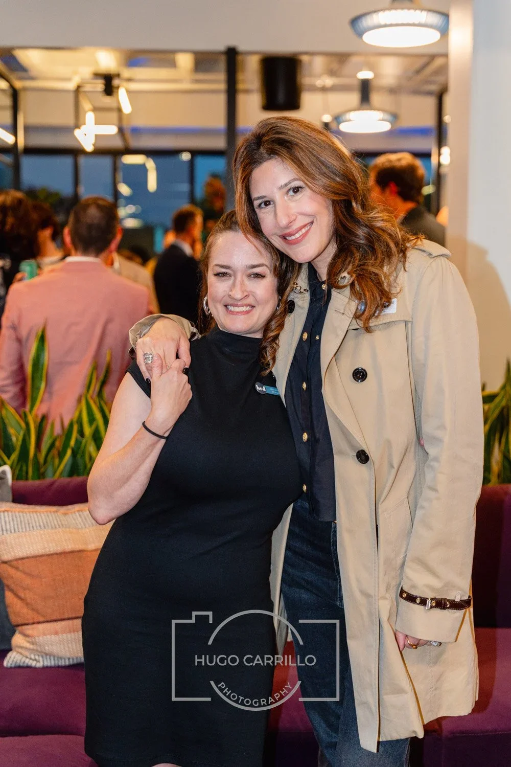 Two women smiling and hugging at a social event, with people in the background and indoor lighting.