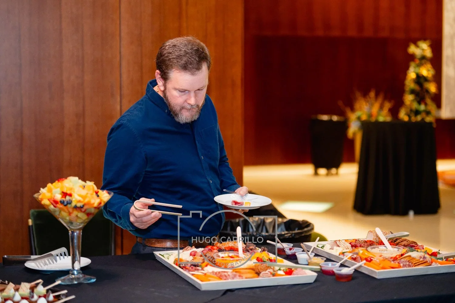 A man with a beard dressed in a blue shirt using chopsticks to serve himself food from catering trays at a buffet table, with fruit salad in a glass bowl and various food items around him.