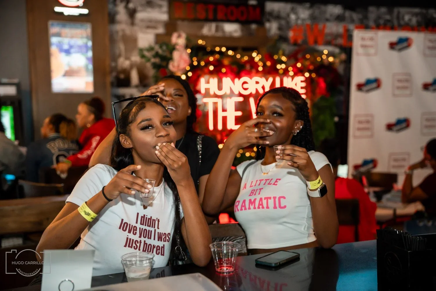 Three young women laughing and sharing drinks at a lively indoor party or bar, with a neon sign and decorative background behind them.