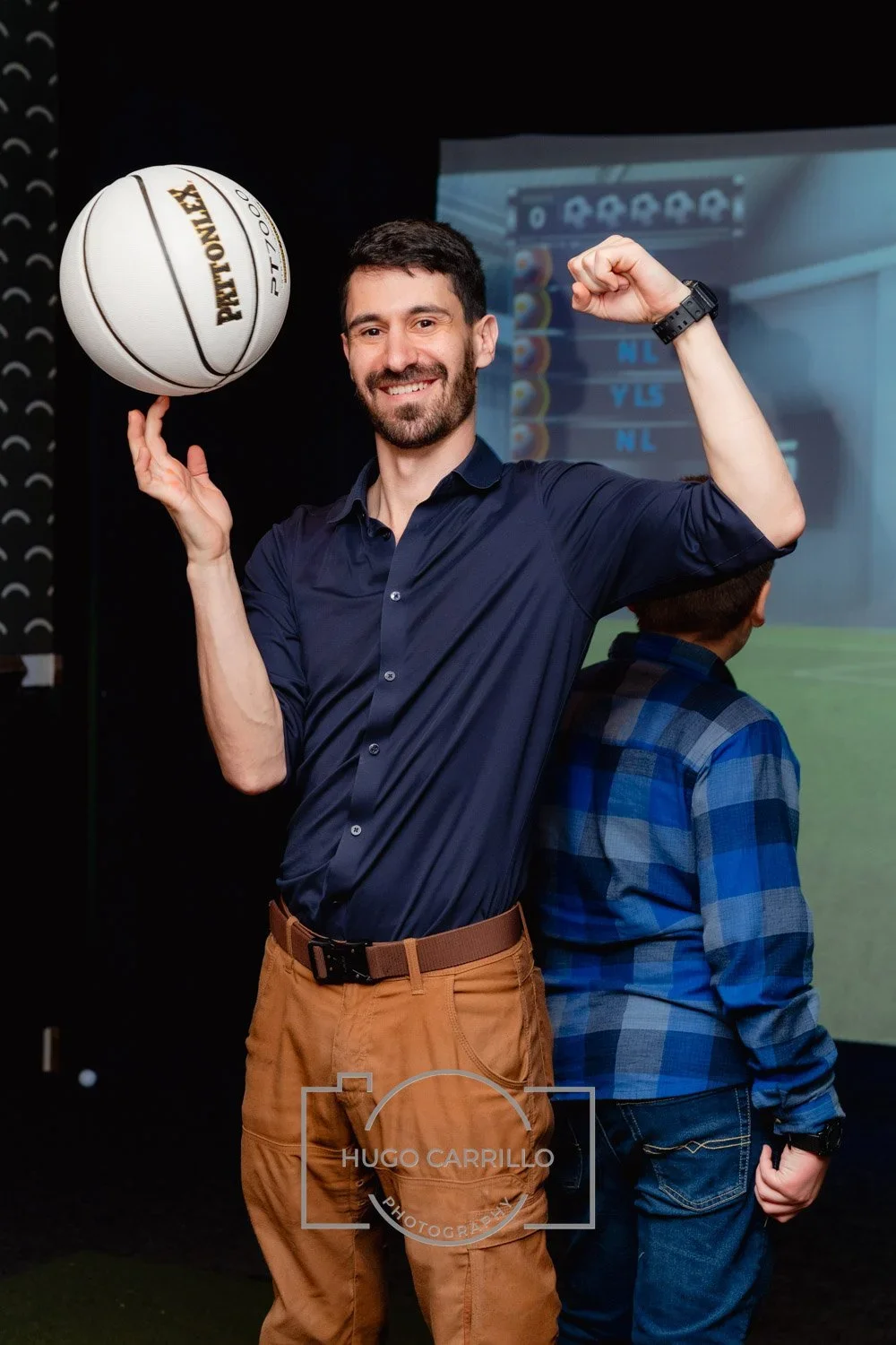 A man smiling and holding a Raymond basketball, flexing his right arm, with a sports bar or entertainment venue screen in the background.