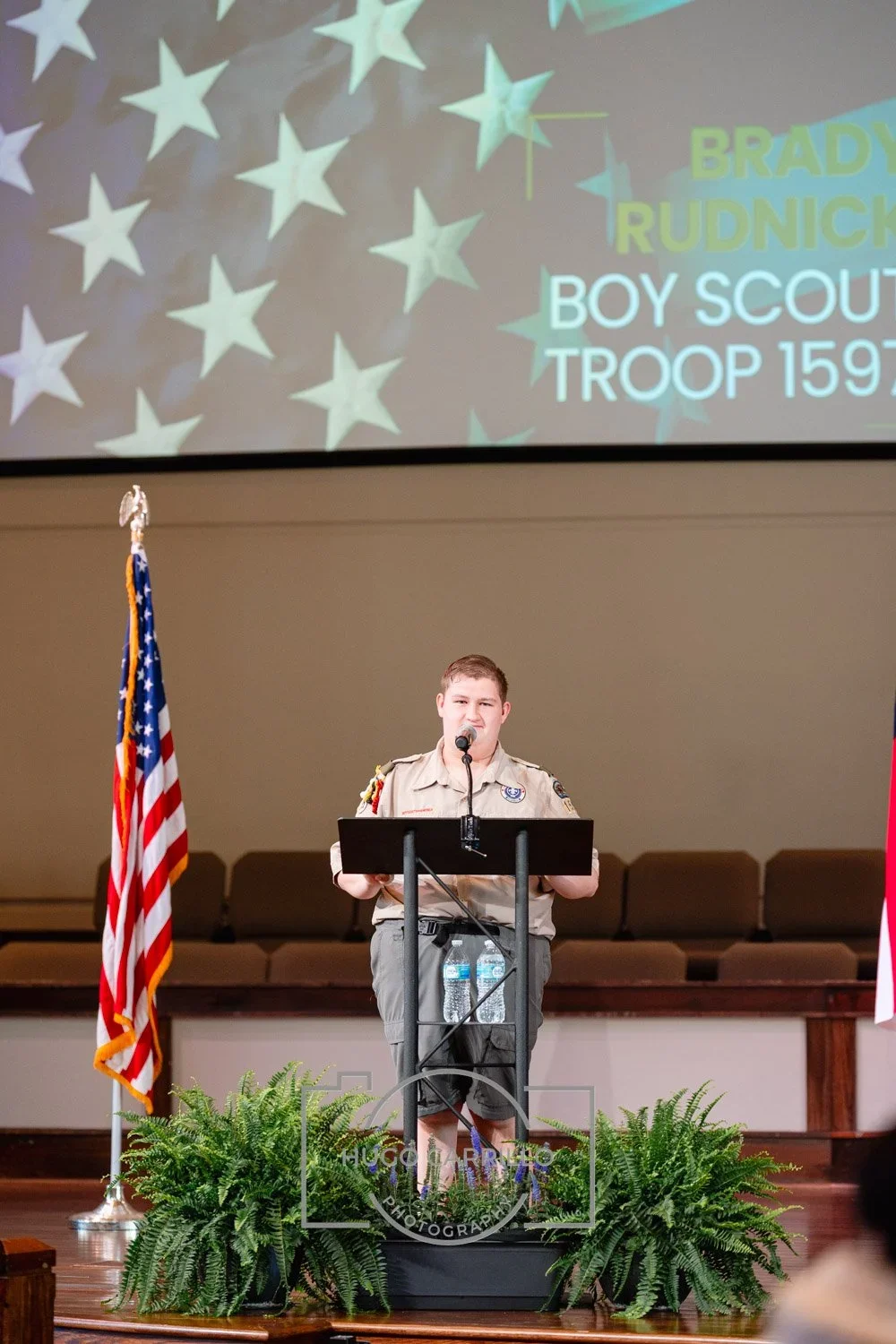 A young Boy Scout stands at a podium on a stage, speaking into a microphone. The stage is decorated with two American flags and green plants at the front. A large screen behind displays a blue star pattern and the text: 'BRADY RUDNICK BOY SCOUT TROOP