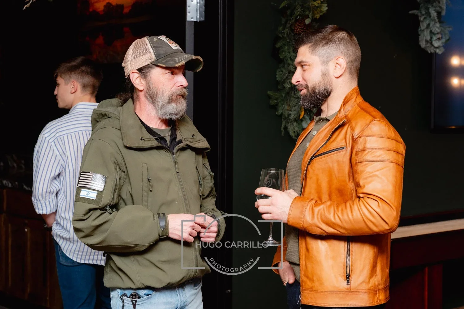 Two men are having a conversation at an indoor event. One man has a gray beard, is wearing a green jacket with patches, and a baseball cap. The other man has a dark beard, is wearing an orange leather jacket, and is holding a glass of dark beverage.