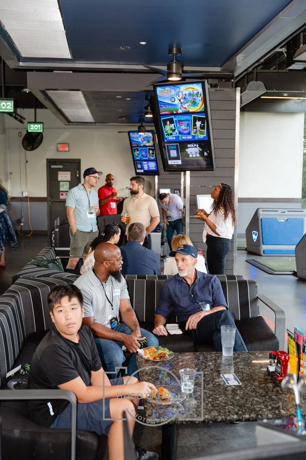 People socializing at a bar or lounge area, with large screens showing a sports game or entertainment, some seated and others standing with drinks in hand.