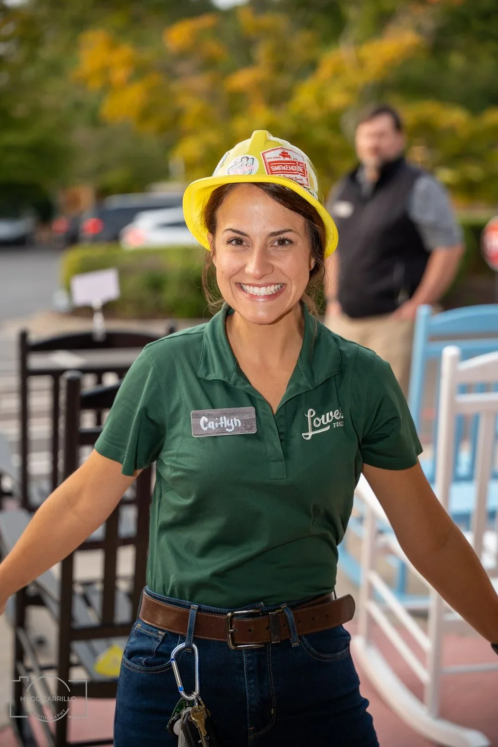 A smiling woman wearing a yellow firefighter helmet and a green shirt with a name tag that reads 'Caitlyn' and 'Lowe's'.
