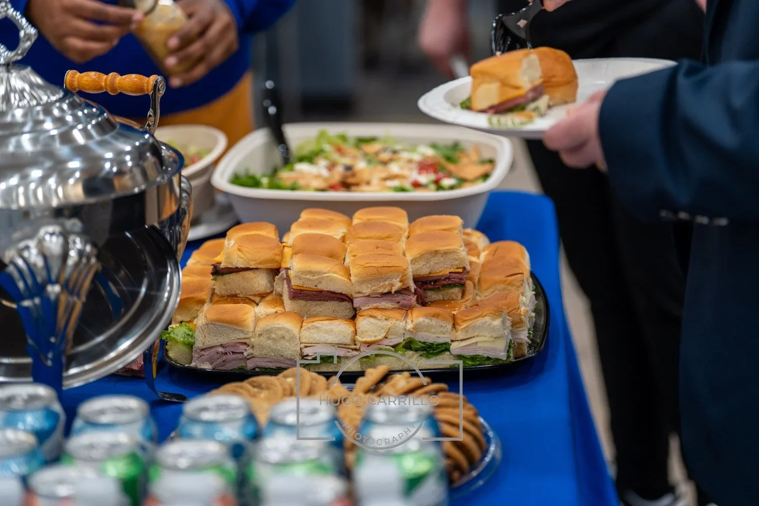 A platter of assorted mini sandwiches with various deli meats, cheese, and lettuce on a blue tablecloth at a buffet. In the background, a bowl of salad, cans of soda, and a person holding a plate with a sandwich. People are serving themselves.