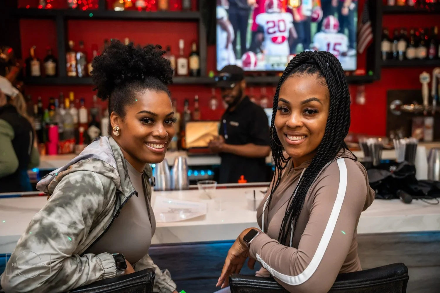Two smiling women with athletic wear sitting at a bar in a sports bar. A man is visible in the background on a phone, and a TV screen shows football players.