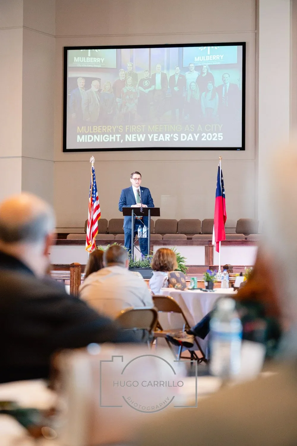 A man in a suit and tie stands at a podium speaking to an audience in a formal meeting room with American and state flags; a large screen behind him displays a group photo with text about Mulberry's first city meeting on New Year's Day 2025.
