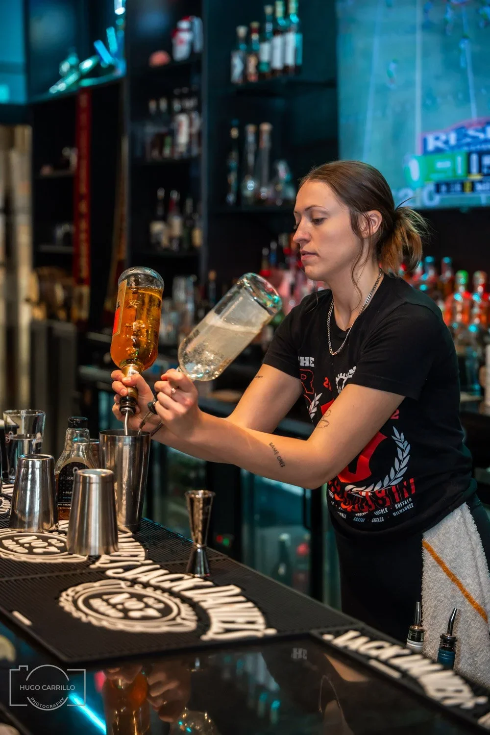 A bartender pours a clear drink from a large glass jar into a metal shaker at a bar with various bottles in the background.