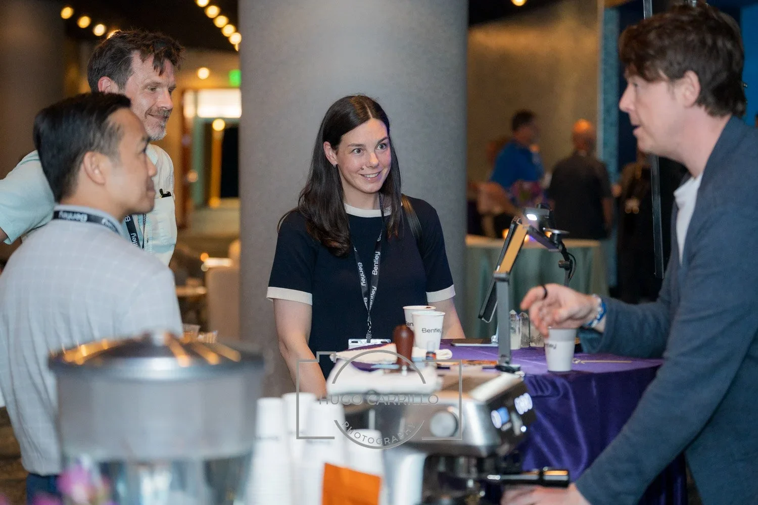 A group of people at a conference or event, engaging in conversation at a booth or registration desk with coffee cups and a coffee maker in the foreground.