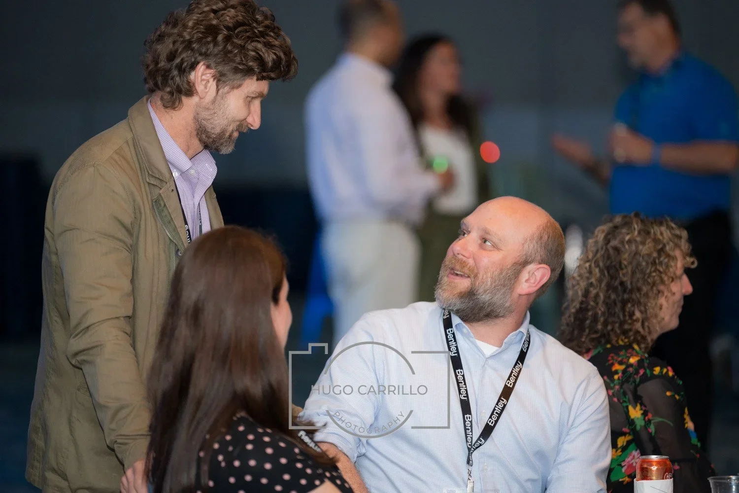 Group of people at a networking event, engaging in conversation, sitting and standing, with a man in a brown jacket talking to a seated man with a beard, wearing a white shirt and lanyard.