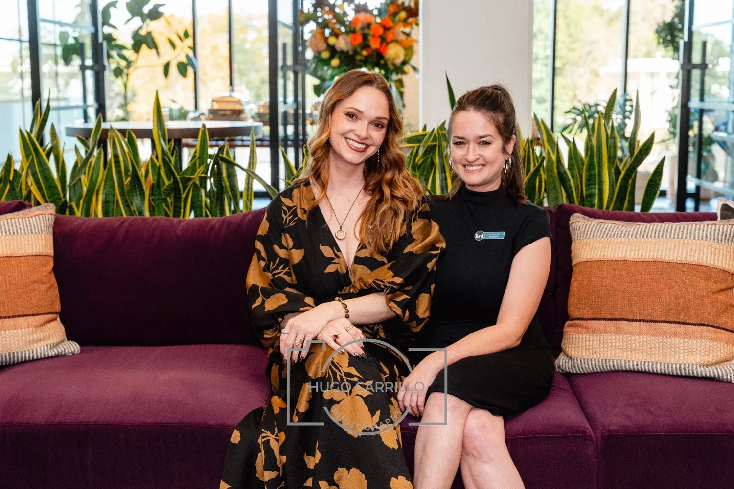 Two women sitting on a purple sofa with green plants and large windows behind them. The woman on the left has long red hair, wearing a black dress with gold leaf patterns, and is smiling with her hands crossed. The woman on the right has dark brown h