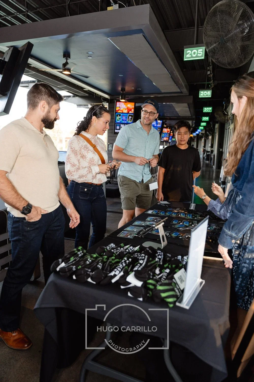 Group of five people standing around a table with merchandise, including socks and digital devices, inside a sports stadium or arena, with digital displays and a large fan overhead.