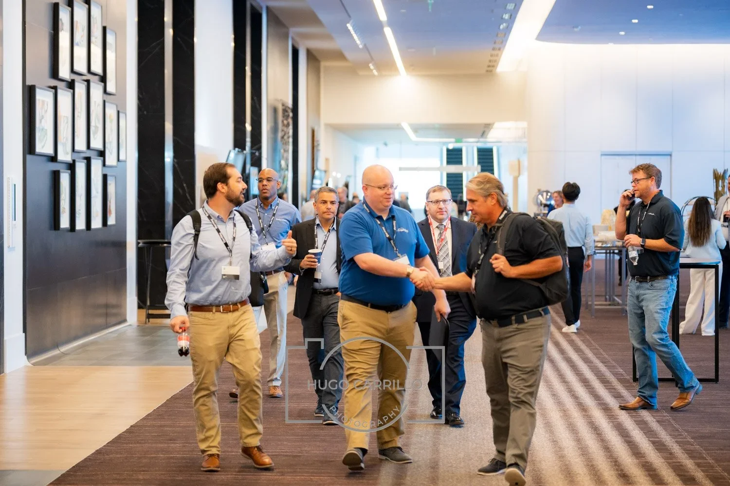 Group of professionals at a conference or networking event; two men in the foreground shaking hands, others walking and talking in the background.