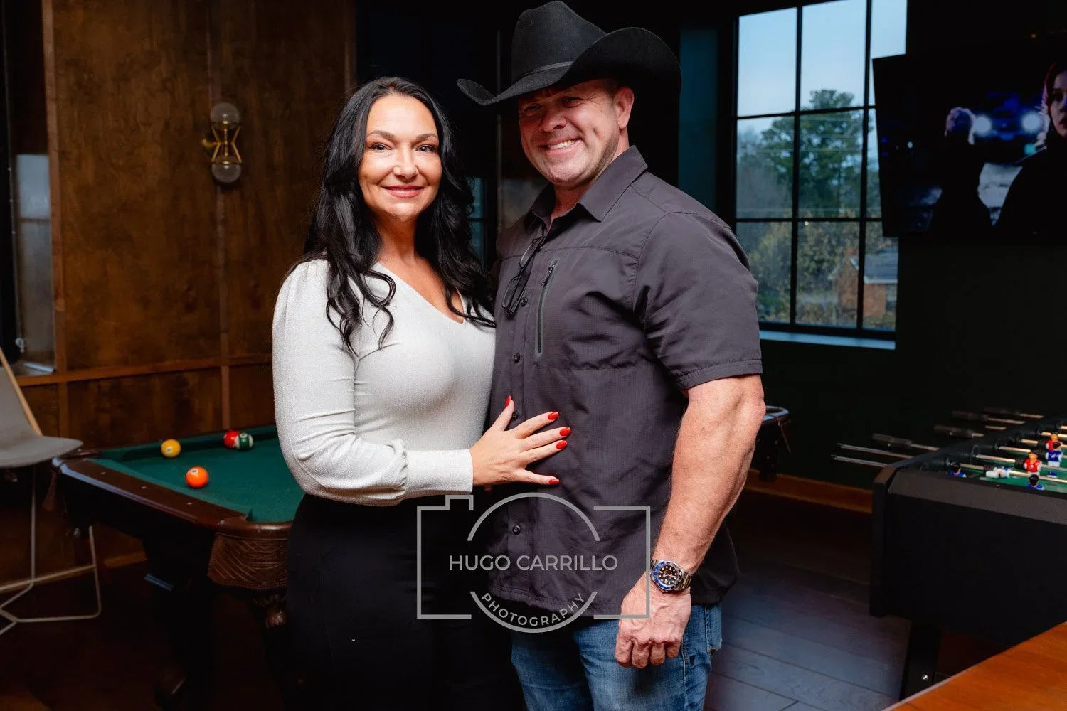A smiling woman with dark hair wearing a white top, and a man in a cowboy hat and gray shirt, standing close together in a room. There is a pool table and a foosball table in the background.