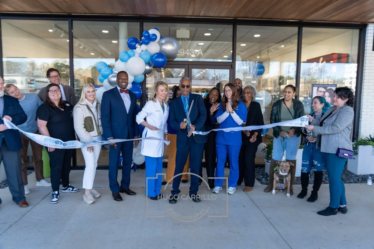 Group of people at a ribbon-cutting ceremony in front of a new building, with balloons and decorations, including healthcare professionals and community members.