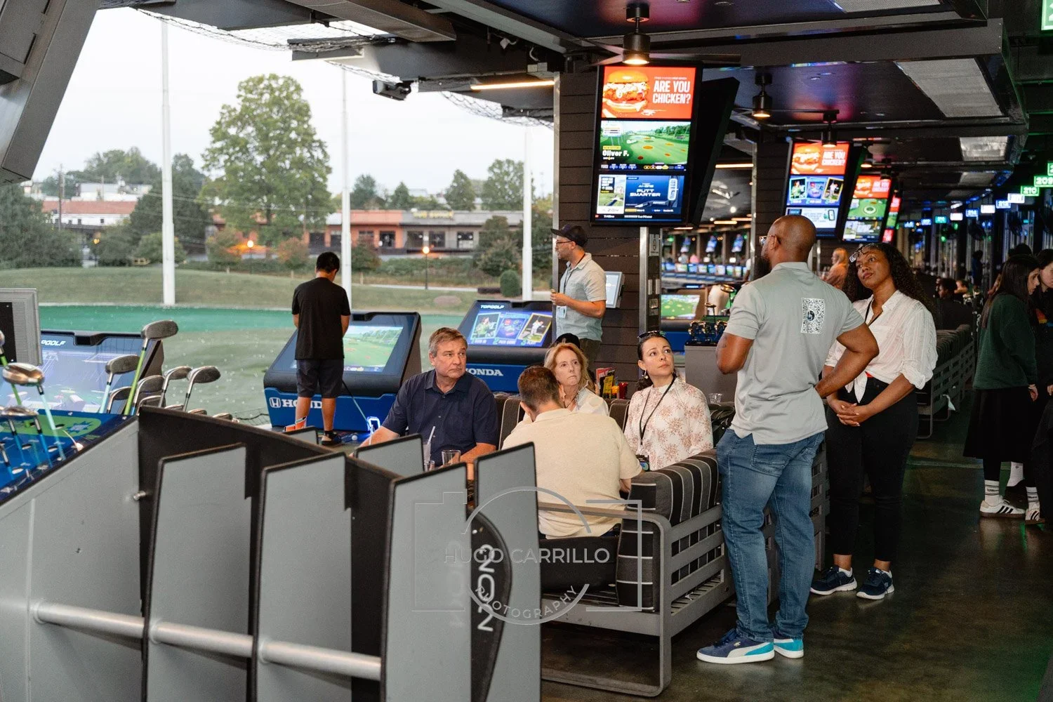 Golf simulation arcade with people sitting on couches, playing and viewing screens, a person standing nearby, and outdoor view through large windows.