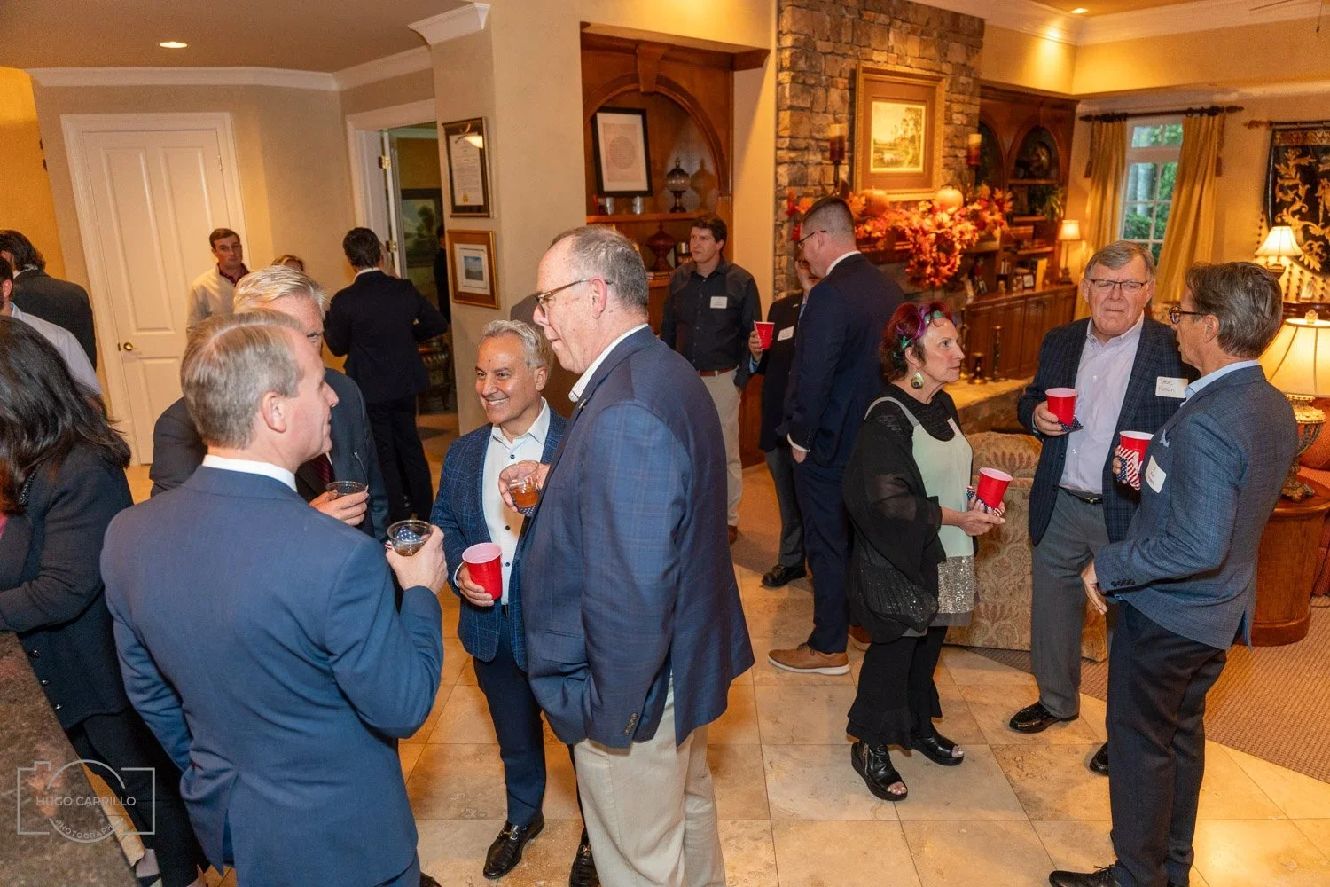 At a social gathering, several people are engaged in conversation, holding drinks, in a warmly decorated indoor setting with framed artwork, a fireplace, and floral arrangements.