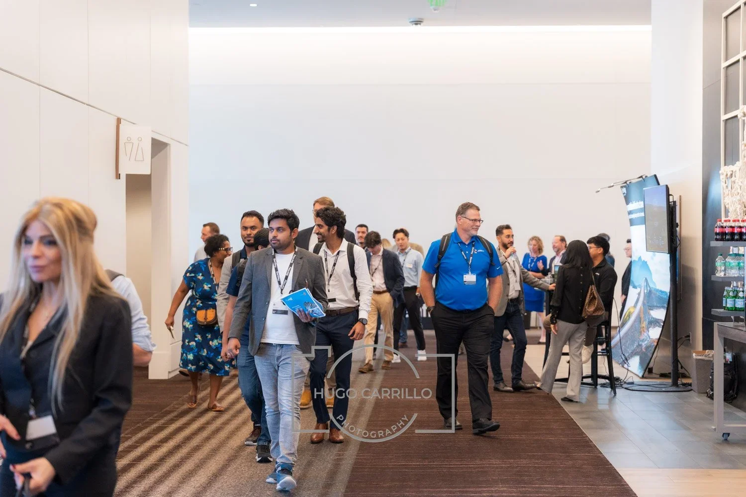 Group of people walking in a conference hall, some carrying badges and conference materials, with a vending area and digital displays on the right side.