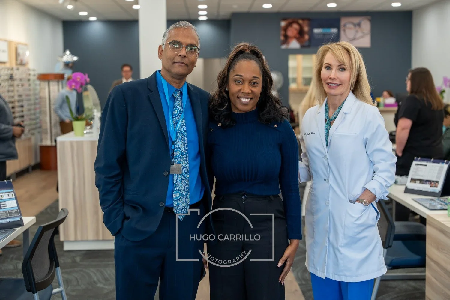 Three professionals in a medical office, two women and one man, smiling and posing together. The man is in a dark blue suit with a patterned blue tie and glasses, the woman in the middle has dark hair in a ponytail and is wearing a navy sweater, and