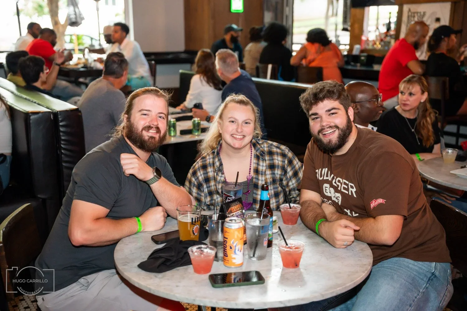 Three friends sitting at a round table in a restaurant, smiling at the camera, with drinks and a phone on the table, with other patrons in the background.