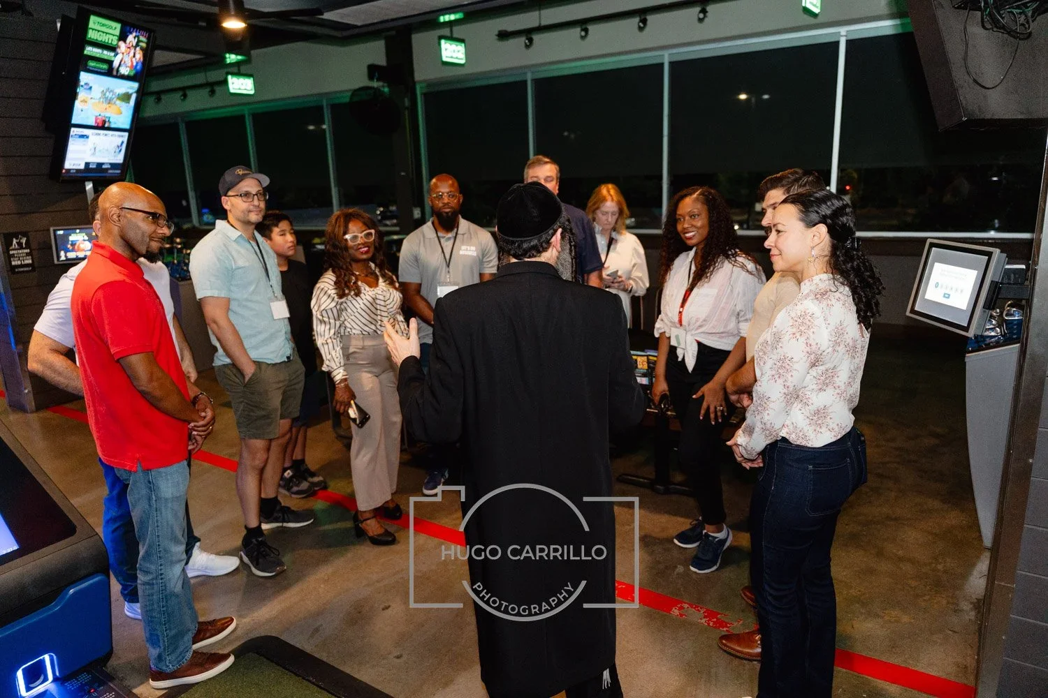 A group of diverse people gathered around a man in a black coat and hat, who appears to be speaking or giving a presentation in a modern indoor space with large windows and electronic screens.
