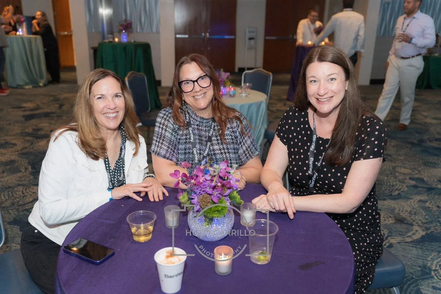 Three women sitting at a round table with purple tablecloth, smiling, during a professional or social event. The table has a purple flower centerpiece and drinks. Other people are seen in the background.