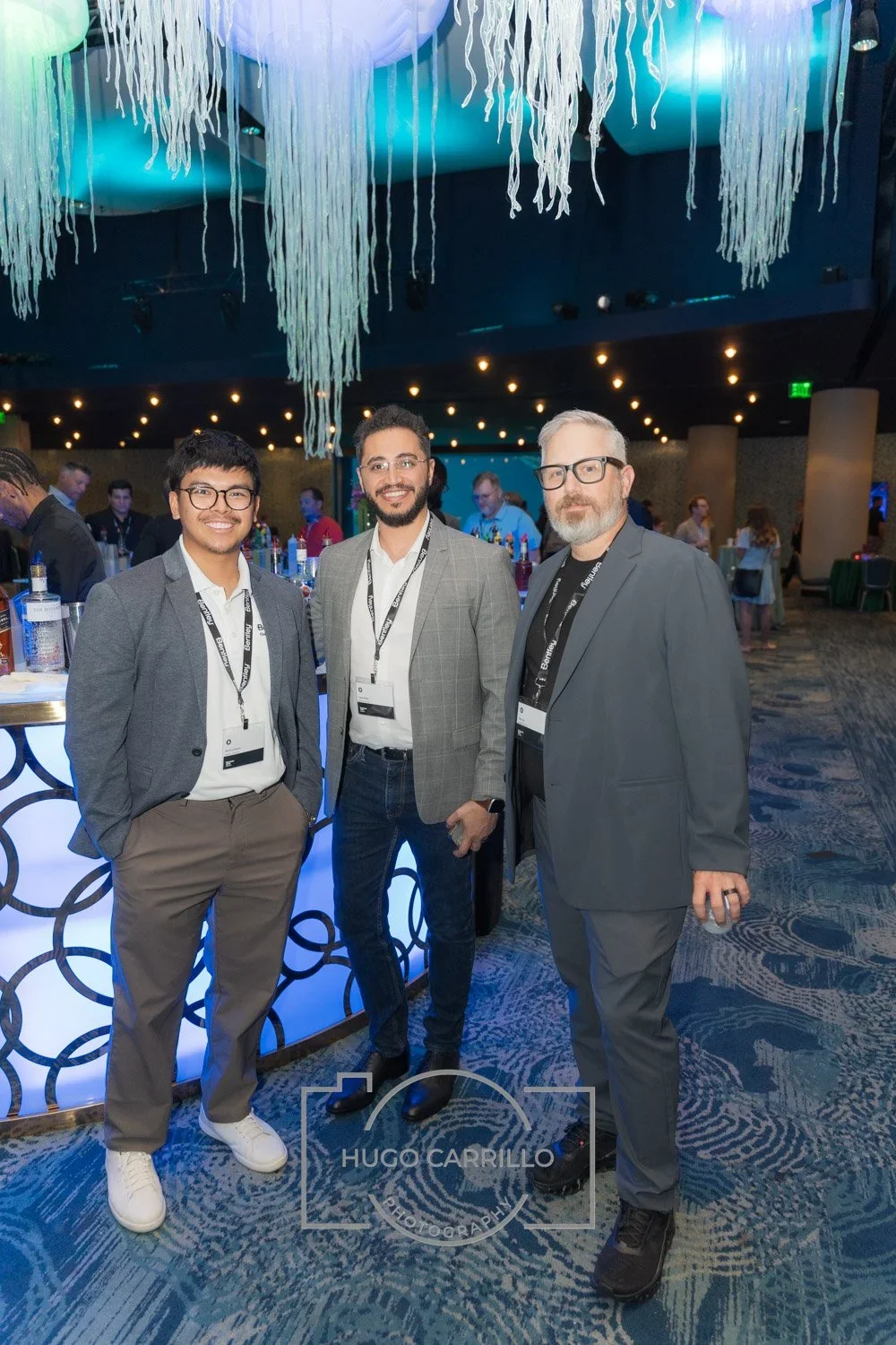Three men dressed in formal/business attire standing together at a conference or networking event, smiling at the camera. The background shows a decorated venue with a bar and other attendees.